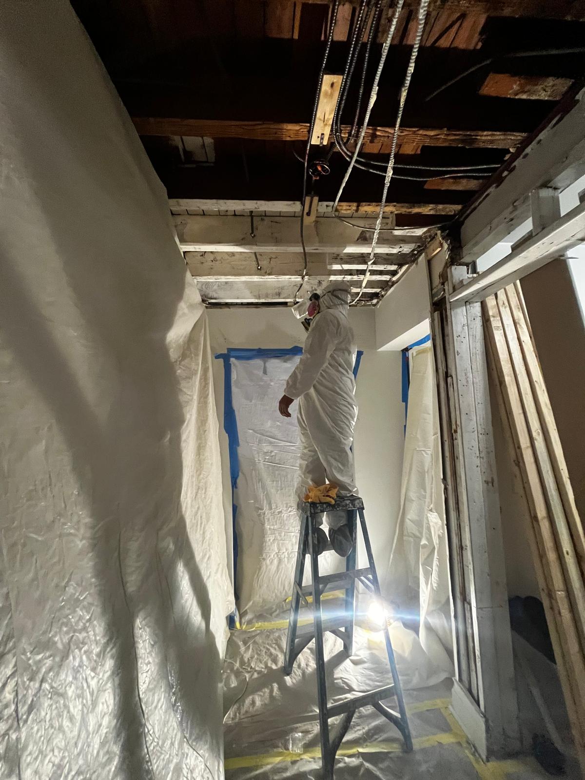 Remediation worker in protective clothing working beneath an opened ceiling inside containment