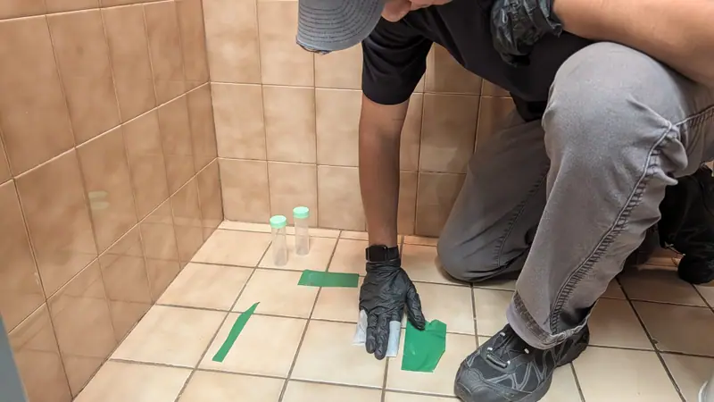 Inspector in black gloves taking a dust wipe clearance sample from a tile floor marked with green tape