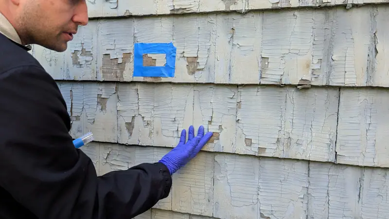 Inspector marking a paint chip sample location with blue tape on peeling exterior siding