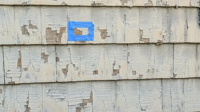 Blue tape marking a paint chip sample location on cracked, peeling exterior paint