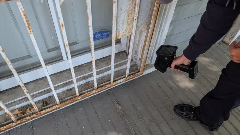 XRF analyzer pointed at a painted window sill behind rusted porch window bars